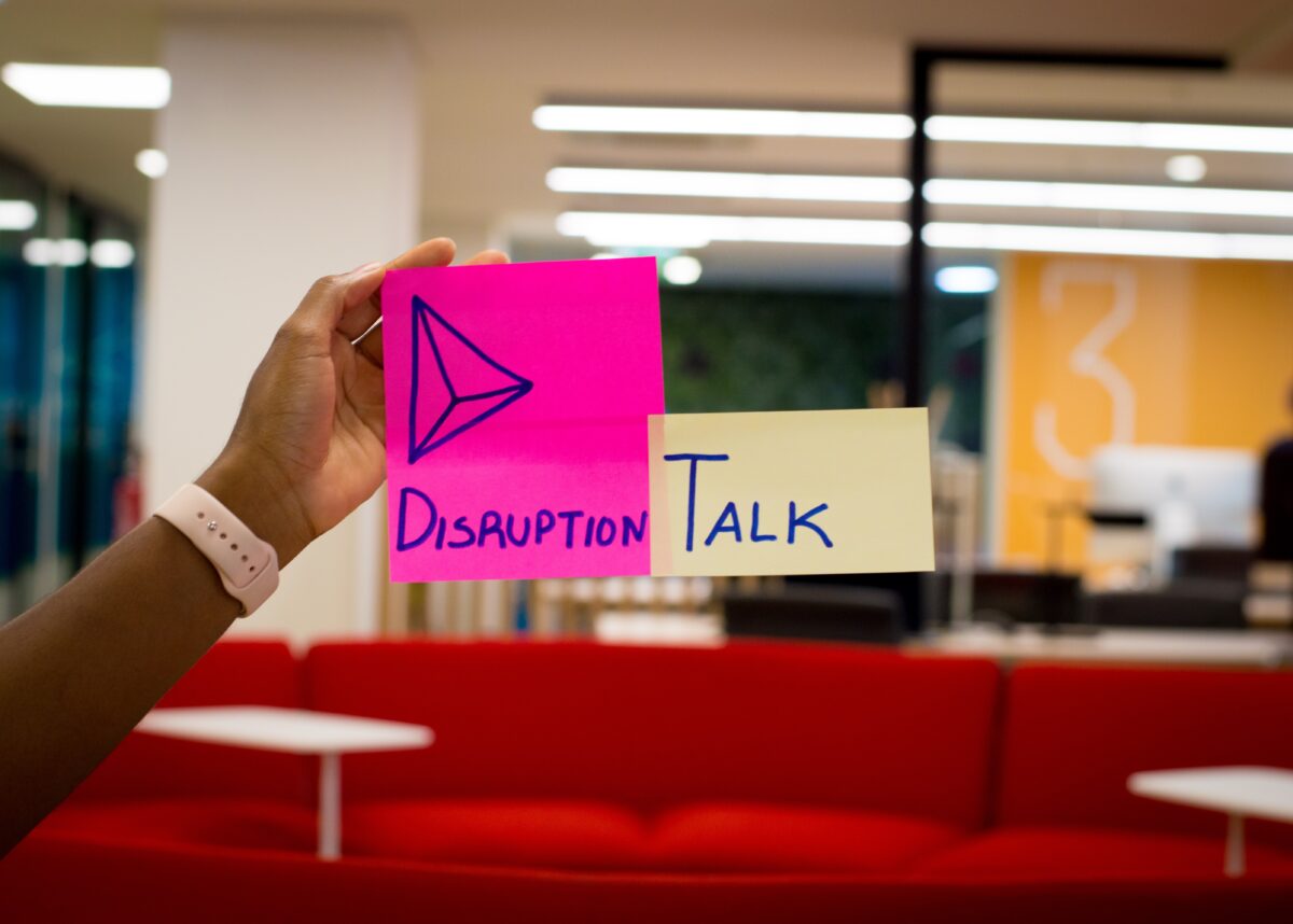 Une main place un post-it sur la fenêtre d'un bureau, avec l'inscription "Disruption-Talk".