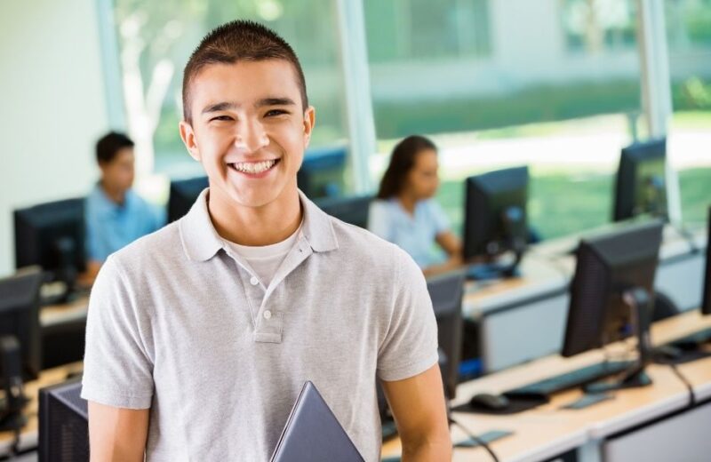 Un jeune homme debout devant une salle informatique, souriant