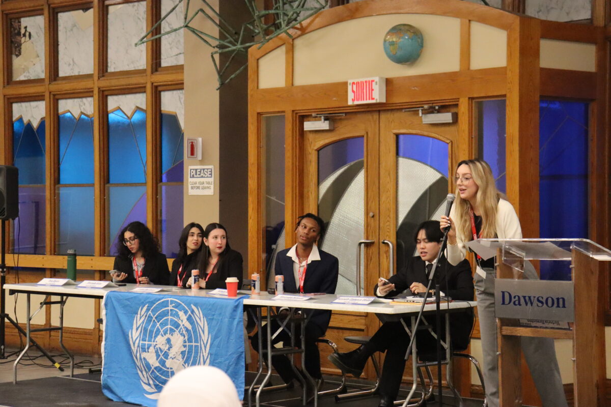 des étudiants assis à une table portant une nappe du Modèle des Nations Unies, avec un étudiant parlant dans un microphone