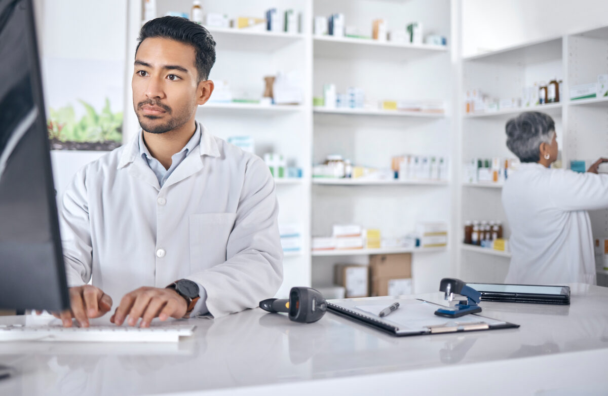 young male pharmacist looking at a computer with shelves of medication behind him