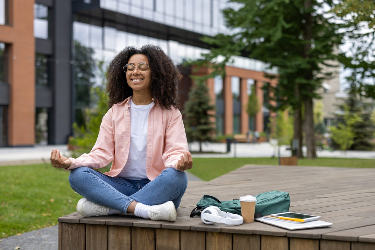 étudiante souriante méditant en plein air, assise les jambes croisées avec un cartable, des livres et une boisson à ses côtés