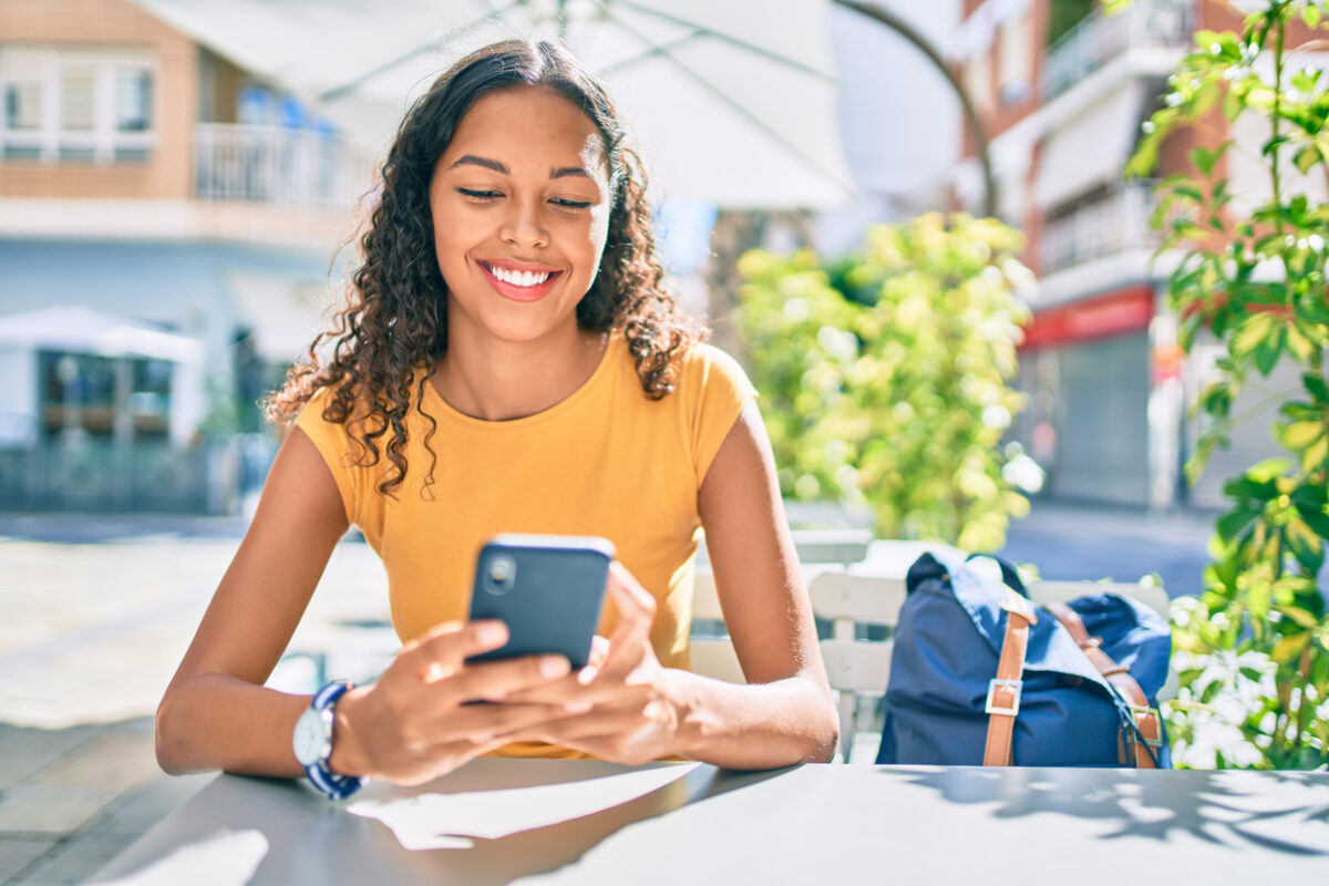 Étudiante souriante assise à l'extérieur par une journée d'été, regardant son téléphone avec son sac à dos à côté d'elle.