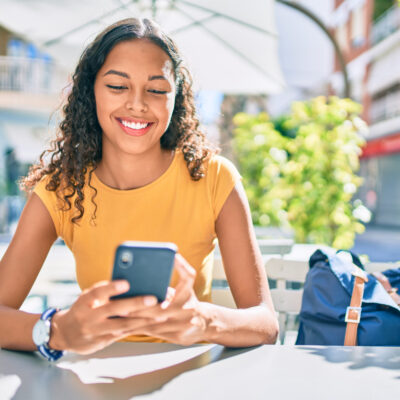 Étudiante souriante assise à l'extérieur par une journée d'été, regardant son téléphone avec son sac à dos à côté d'elle.
