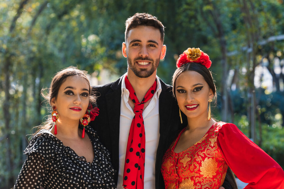 Trois danseuses de flamenco souriantes face à la caméra dans un parc par une journée ensoleillée.