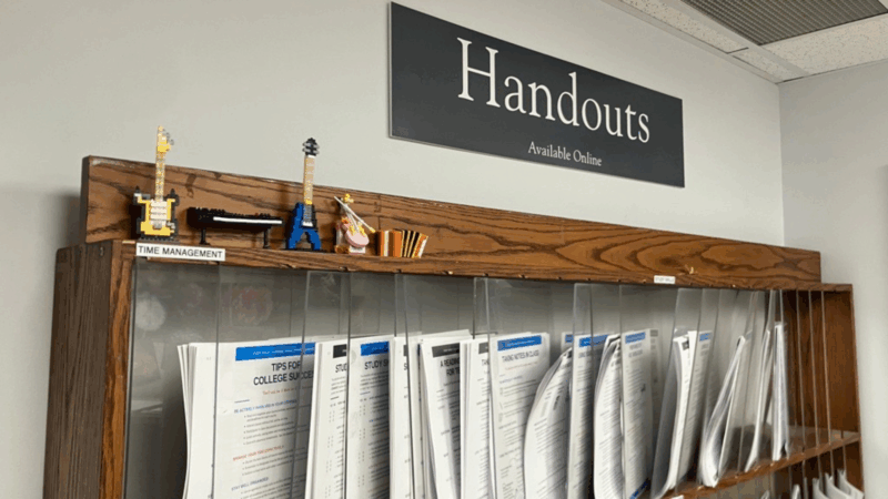 Photo d'une structure en bois servant à afficher des documents, séparés par du plexiglas transparent. Le mot « Handouts » (documents) est inscrit sur un panneau bleu-gris au-dessus de la structure.